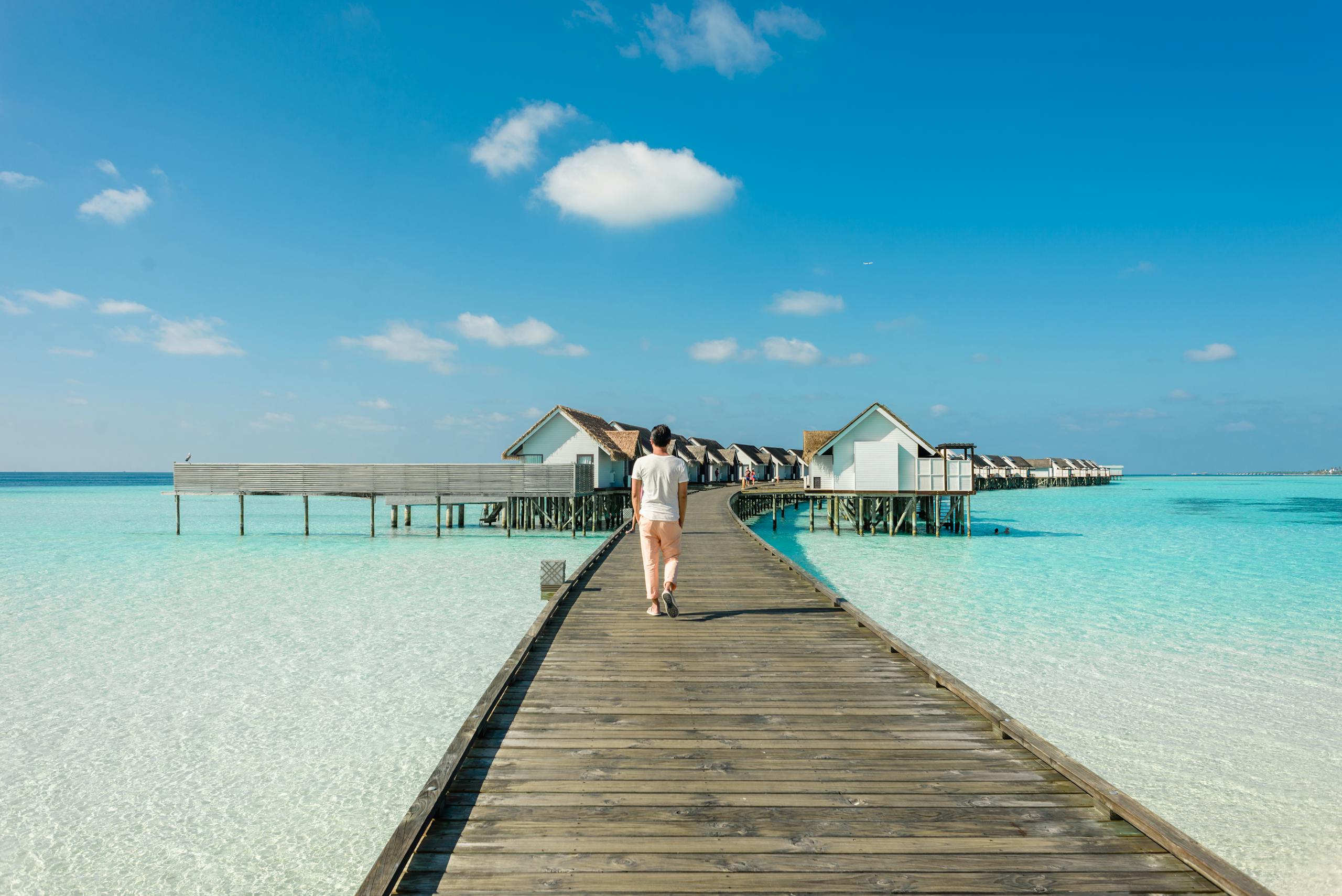 Man walking on a wooden dock towards luxurious ocean bungalows under clear blue sky.